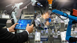 Ein Ingenieur hält ein Tablet in der Hand und kontrolliert damit zwei Arme eines Roboters um Teile einer Produktion herzustellen.