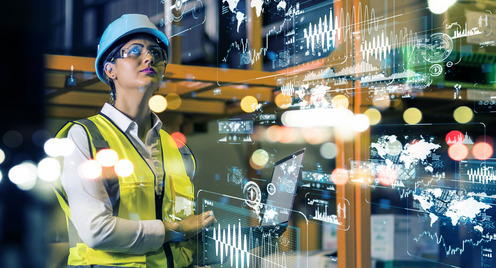 Eine junge Frau steht in einer Fabrikhalle, trägt Helm und gelbe Warnweste. Sie steht in einem Hologramm aus Lichtreflexen und Daten.