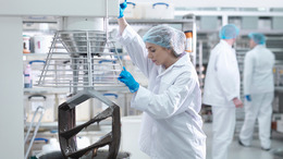 In einer Fabrikhalle arbeitet eine Frau mit Kittel, Haube und Handschuhen bekleidet an einer großen metallfarbenen Maschine zur Lebensmittelproduktion