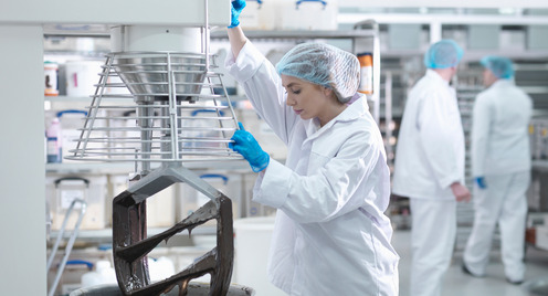 In einer Fabrikhalle arbeitet eine Frau mit Kittel, Haube und Handschuhen bekleidet an einer großen metallfarbenen Maschine zur Lebensmittelproduktion
